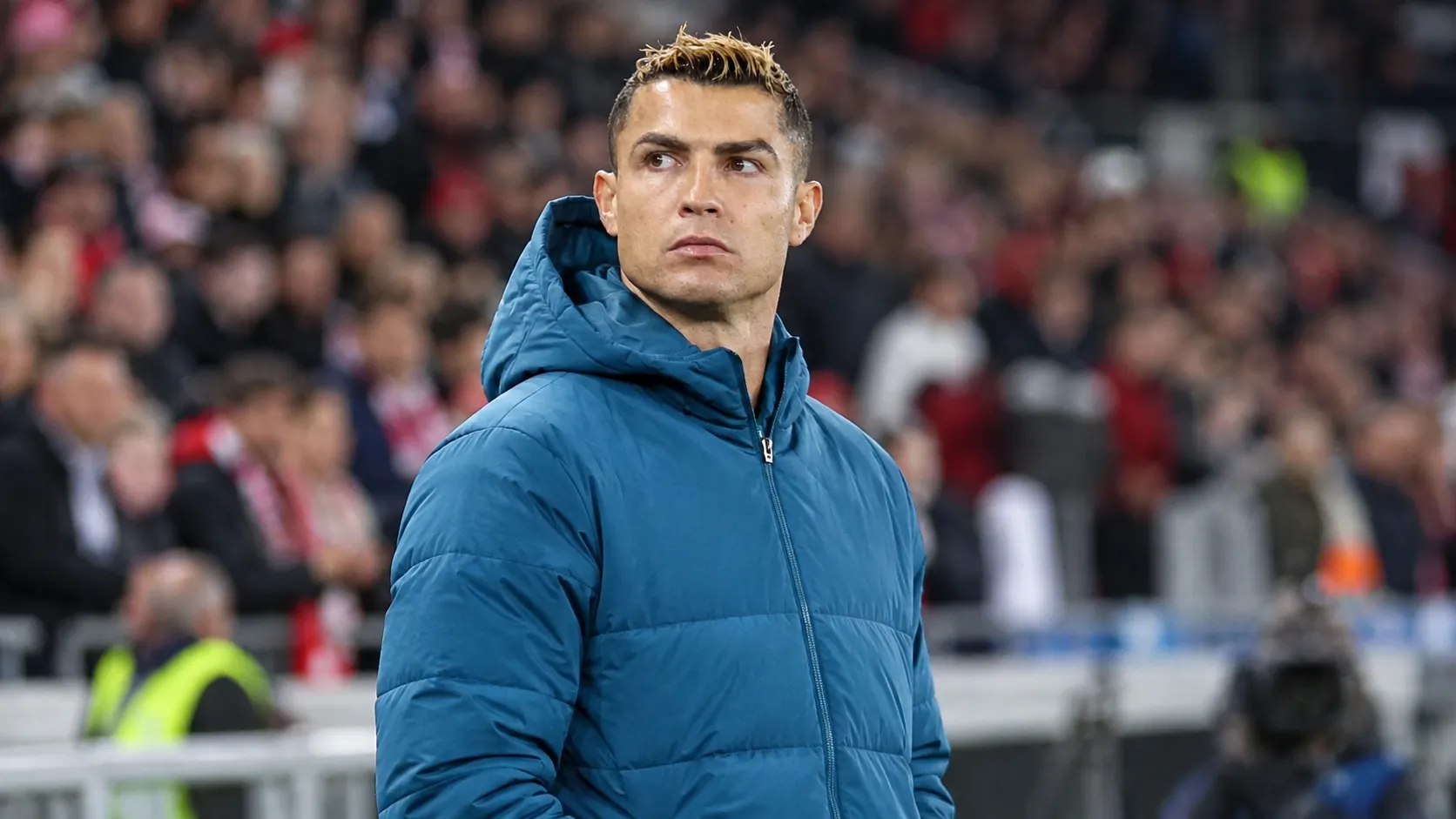 Cristiano Ronaldo in een blauwe jas langs de zijlijn van een voetbalstadion.