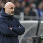 Een coach in een donkere jas langs de zijlijn van een voetbalstadion.