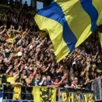 Een volle groep STVV-supporters met geel-blauwe vlaggen en sjaals in de tribune.