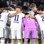 Een groep Anderlecht-spelers vormt een hechte kring op het veld in witte wedstrijdtenues.