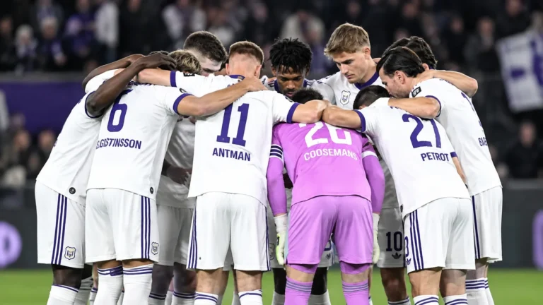 Een groep Anderlecht-spelers vormt een hechte kring op het veld in witte wedstrijdtenues.