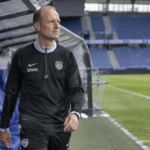 Jess Thorup in een donkere trainingsoutfit naast een dug-out in een voetbalstadion.