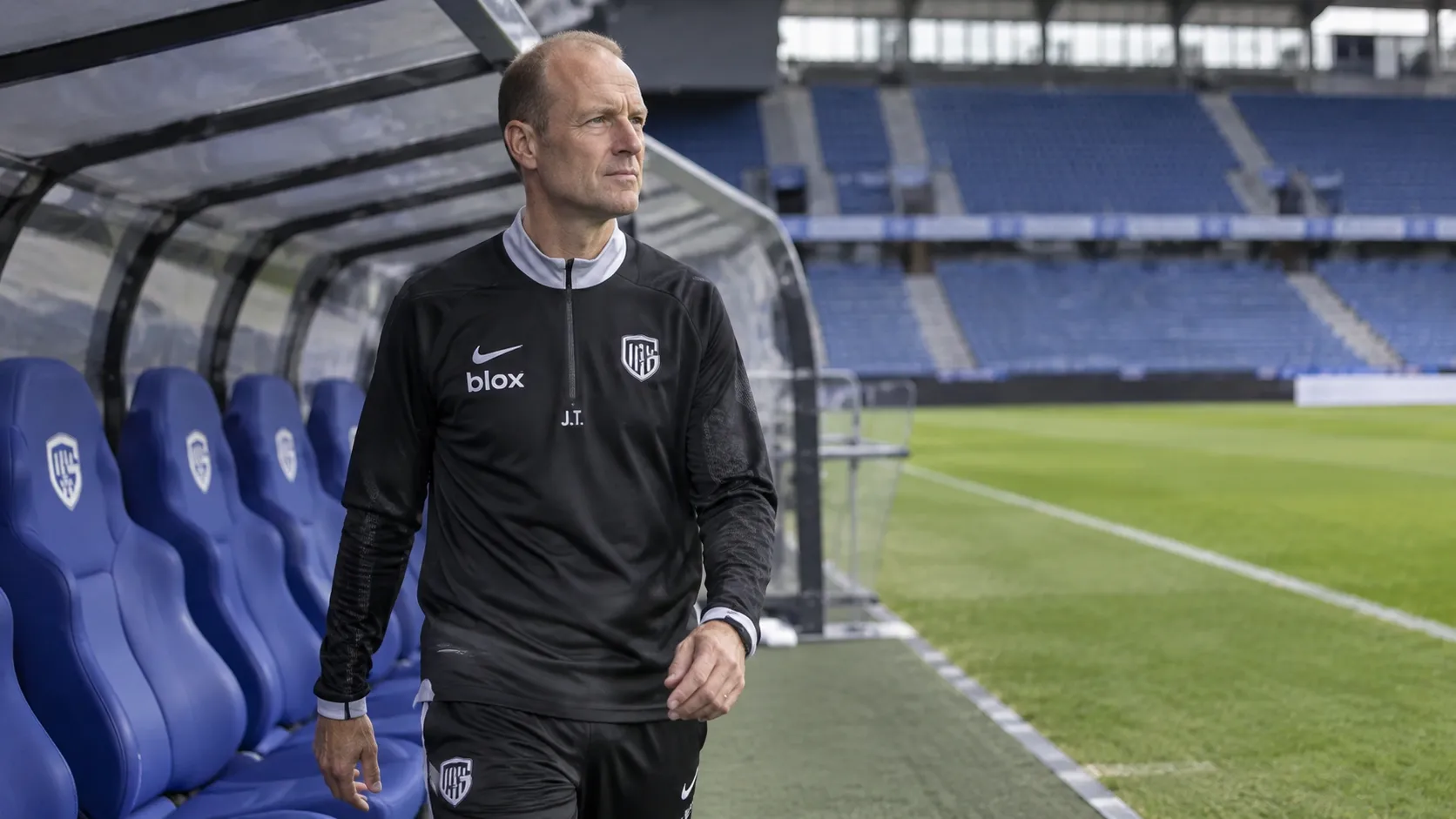 Jess Thorup in een donkere trainingsoutfit naast een dug-out in een voetbalstadion.