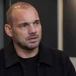 Wesley Sneijder spreekt in een voetbalmediaomgeving met een subtiel KNVB-embleem op de wand.