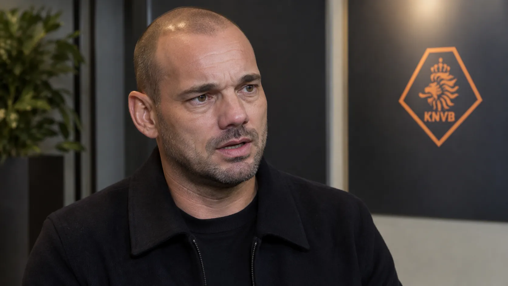 Wesley Sneijder spreekt in een voetbalmediaomgeving met een subtiel KNVB-embleem op de wand.