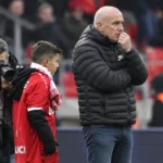 Een man in donkere jas kijkt naast een jongen in rood sportshirt langs het veld toe.