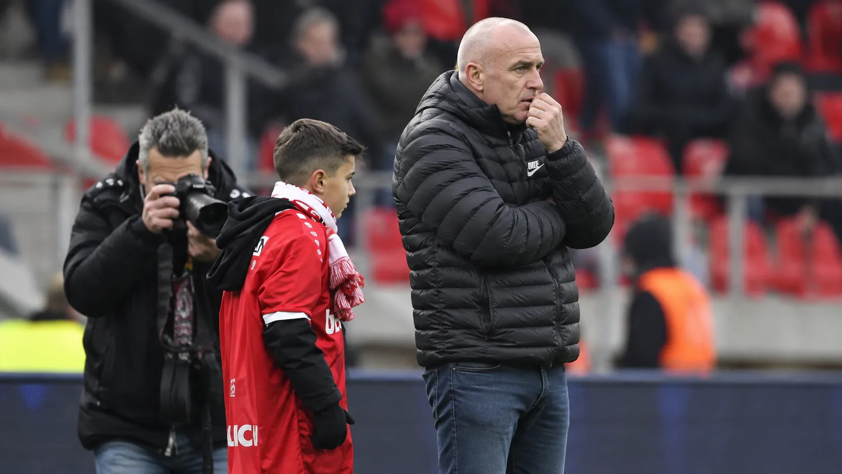Een man in donkere jas kijkt naast een jongen in rood sportshirt langs het veld toe.