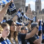Een groep Club Brugge-supporters heft blauw-zwarte sjaals omhoog in een buitenomgeving.