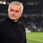 Een grijs­harige trainer in een donkere trui langs de zijlijn van een stadion.