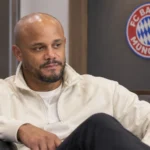Vincent Kompany zit in een rustige ruimte van Bayern München met een clubembleem op de wand.