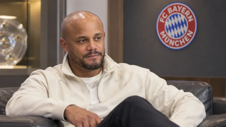 Vincent Kompany zit in een rustige ruimte van Bayern München met een clubembleem op de wand.