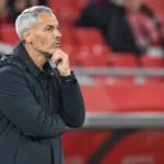 Een coach in donkere jas langs de zijlijn voor rode stadionzitjes.