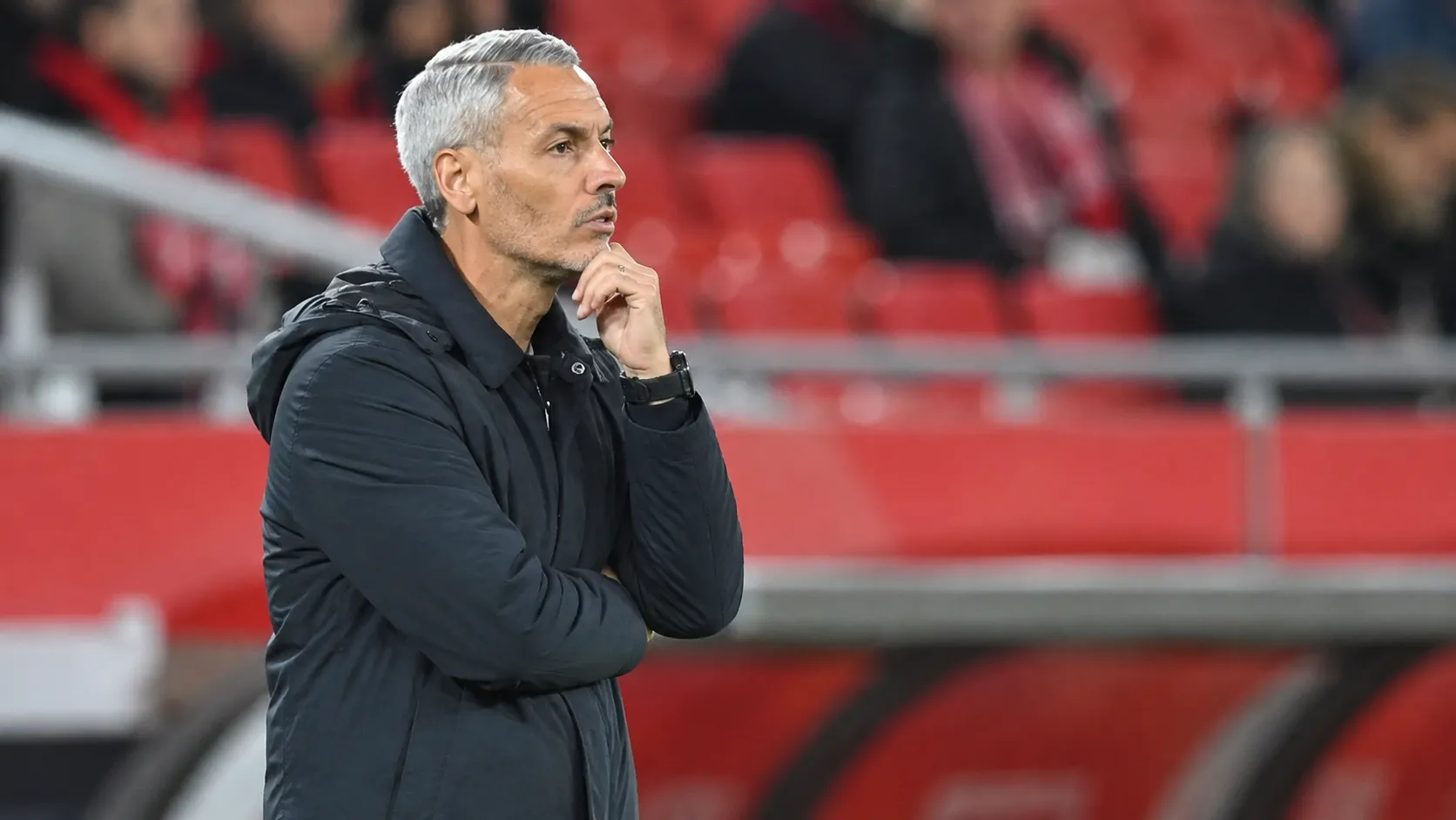Een coach in donkere jas langs de zijlijn voor rode stadionzitjes.