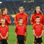 Belgische speelsters in rode trainingsjassen staan met jonge mascottes op het veld voor een interland.