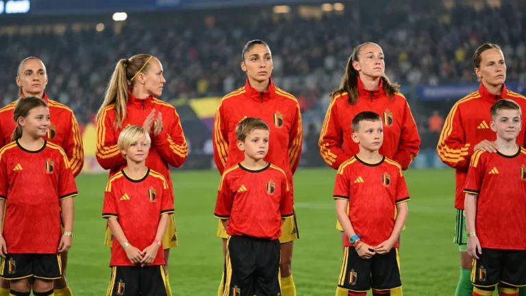 Belgische speelsters in rode trainingsjassen staan met jonge mascottes op het veld voor een interland.