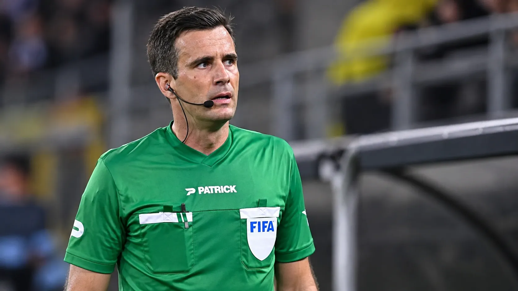 Een scheidsrechter in groen tenue met headset staat langs de rand van het veld.
