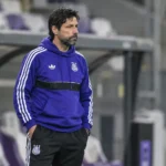 Een staflid in paarse clubkleding staat in de technische zone van een voetbalstadion.