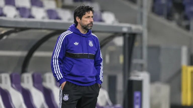 Een staflid in paarse clubkleding staat in de technische zone van een voetbalstadion.