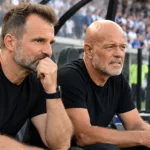 Twee leden van de technische staf bij een voetbaldug-out met supporters op de achtergrond.