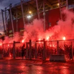 Collectieve supporters bij een stadioningang met brandende fakkels en rode rook.