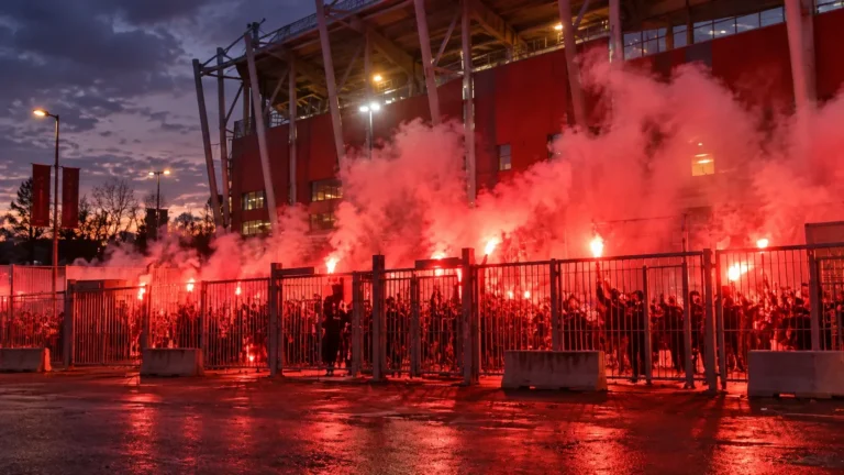 Collectieve supporters bij een stadioningang met brandende fakkels en rode rook.