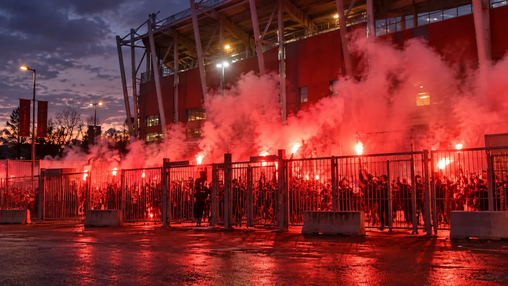 Collectieve supporters bij een stadioningang met brandende fakkels en rode rook.