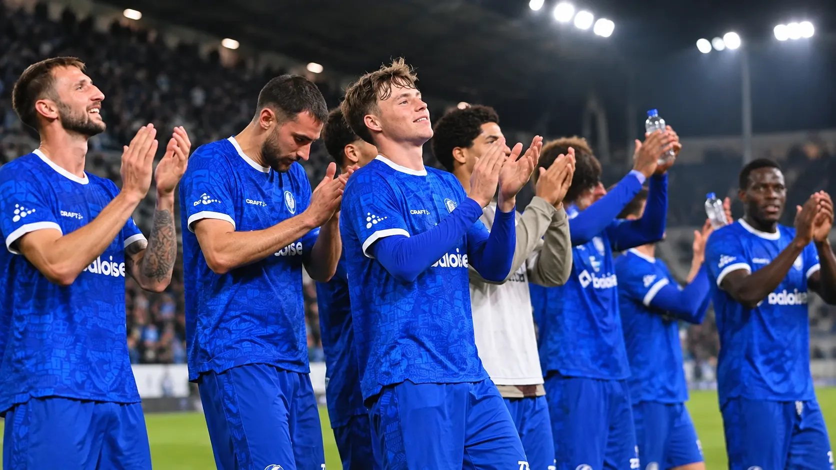 Een groep KAA Gent-spelers in blauwe shirts applaudisseert samen langs het veld.