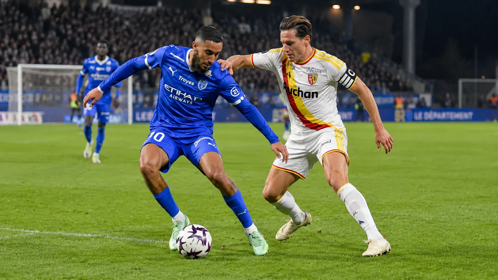 Een speler van RC Lens in duel met een speler van Club Brugge tijdens een avondwedstrijd.
