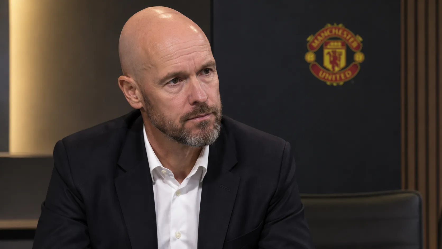 Erik ten Hag tijdens een rustig media-interview met een Manchester United-embleem op de achtergrond.