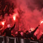 Groep supporters in donkere kledij op een tribune tussen rode rook en fakkels.