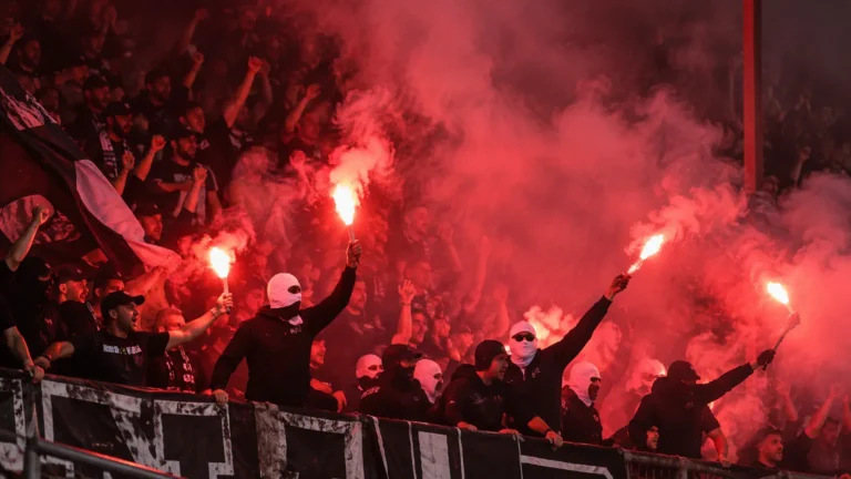 Groep supporters in donkere kledij op een tribune tussen rode rook en fakkels.