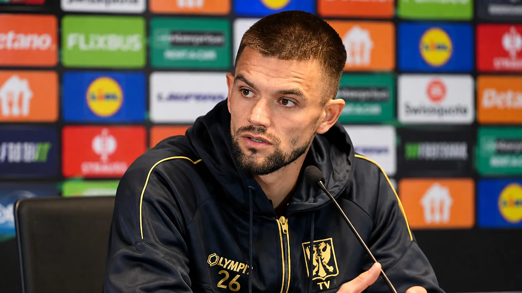 Visar Musliu van STVV spreekt tijdens een persmoment in een donkere clubjas.
