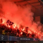 Supporters in een overdekte voetbaltribune tussen rood licht en rook van fakkels.