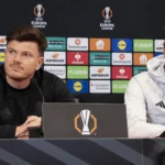 Twee leden van de technische staf aan een persconferentietafel voor een Europa League-achtergrond.