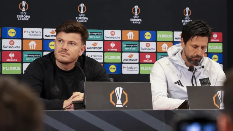 Twee leden van de technische staf aan een persconferentietafel voor een Europa League-achtergrond.