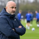 Een niet-spelende clubvertegenwoordiger in een donkere jas aan de rand van een trainingsveld.