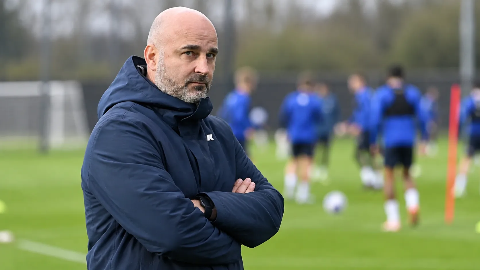 Een niet-spelende clubvertegenwoordiger in een donkere jas aan de rand van een trainingsveld.