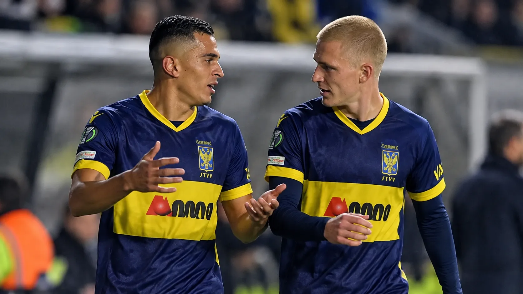Twee voetballers in donkerblauwe shirts met gele details overleggen samen op het veld.