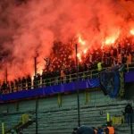 Een collectieve supportersgroep op de tribune met vuurwerk en rook boven het vak.
