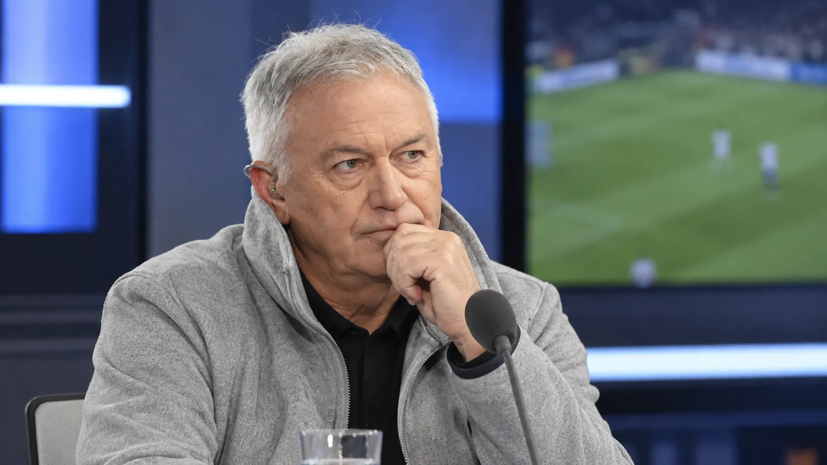 Een grijze voetbalanalist aan een microfoon in een televisiestudio.