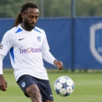 Dieumerci Ndongala in een wit KRC Genk-shirt op een voetbalveld.