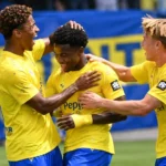 Drie voetballers in geel-blauwe tenues vieren samen op het veld.