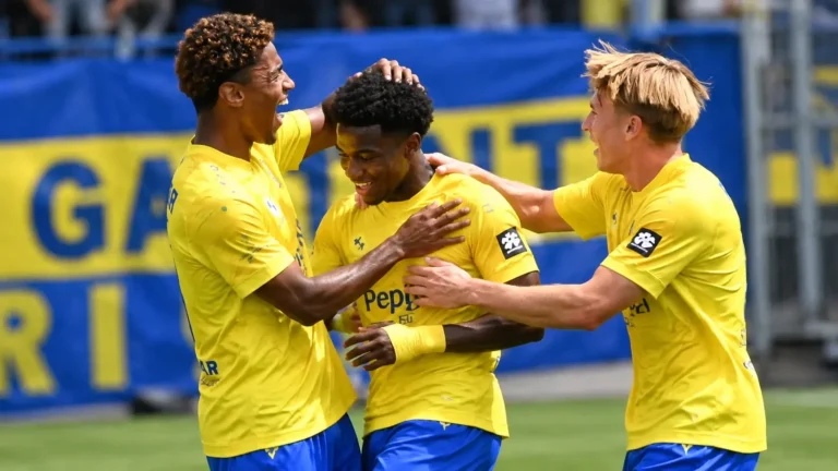 Drie voetballers in geel-blauwe tenues vieren samen op het veld.