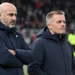 Twee niet-spelende clubvertegenwoordigers in donkere jassen aan de rand van een stadionveld.