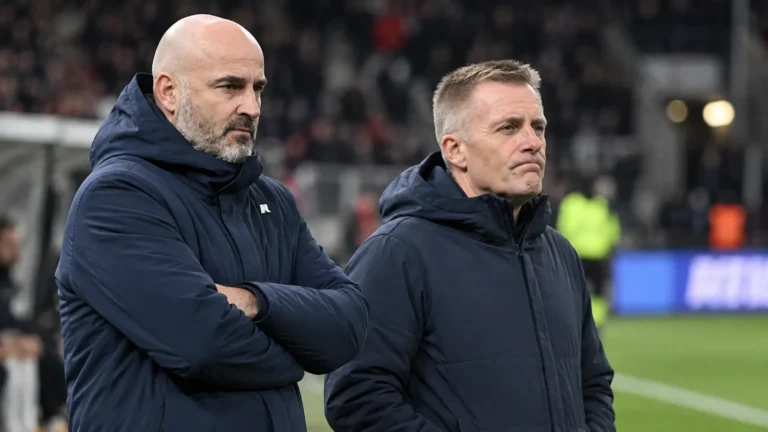 Twee niet-spelende clubvertegenwoordigers in donkere jassen aan de rand van een stadionveld.
