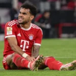Luis Díaz zit op het gras in het Bayern München-tenue en kijkt richting de dug-out.