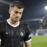 Een teleurgestelde voetballer in een donker wedstrijdshirt naast het veld na een Europese wedstrijd.