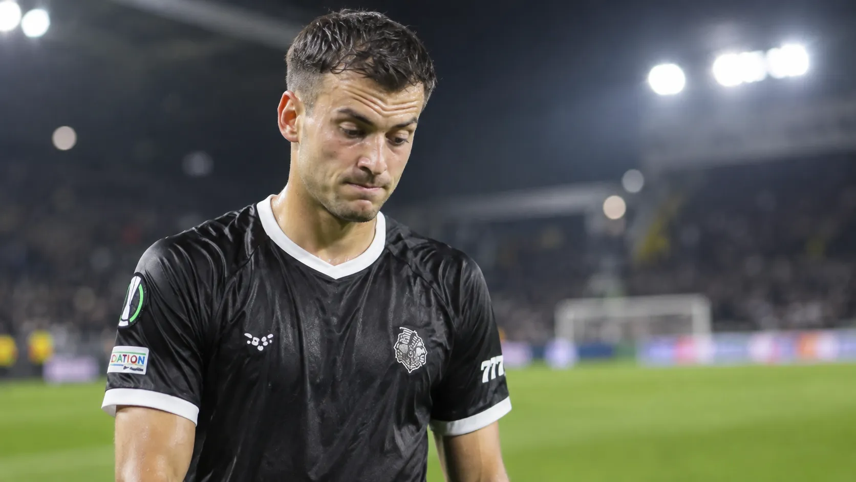 Een teleurgestelde voetballer in een donker wedstrijdshirt naast het veld na een Europese wedstrijd.