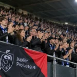 Een collectieve groep Jupiler Pro League-supporters applaudisseert in een verlicht Europees stadion.
