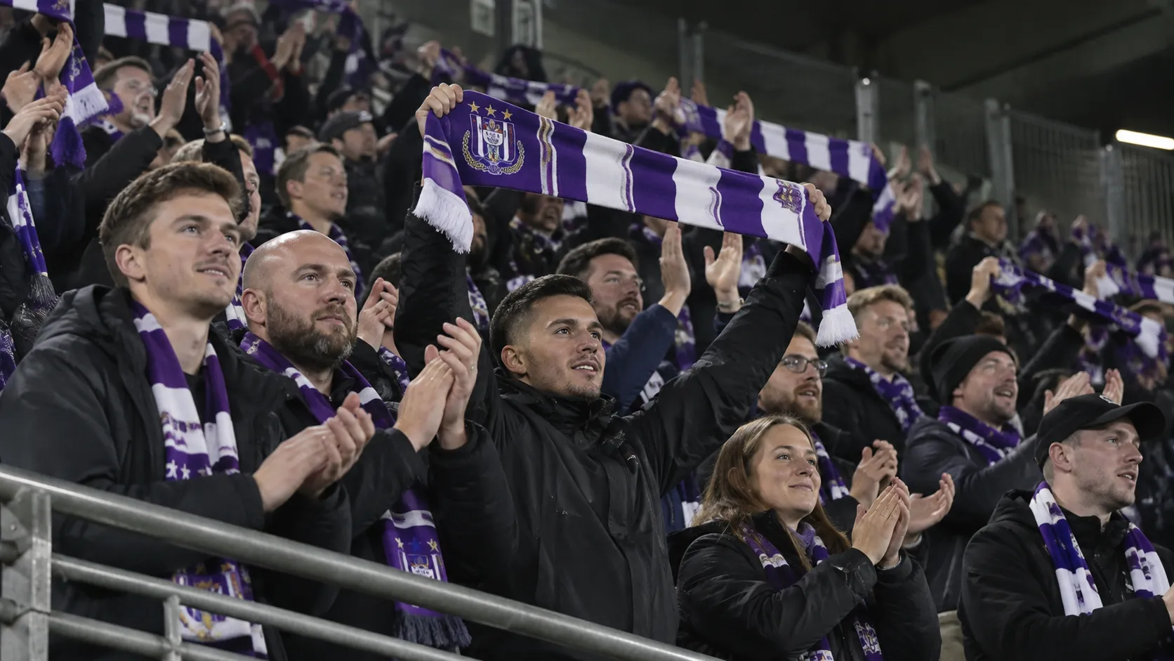 Een groep Anderlecht-supporters viert samen in de tribune met paarse en witte sjaals.